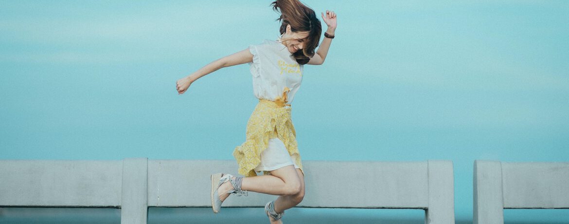 Een vrouw met een gele rok en witte blouse en lang bruin haar springt in de lucht, achter haar zie je een reling zee. Foto via pexels door: Quang Anh Ha Nguyen