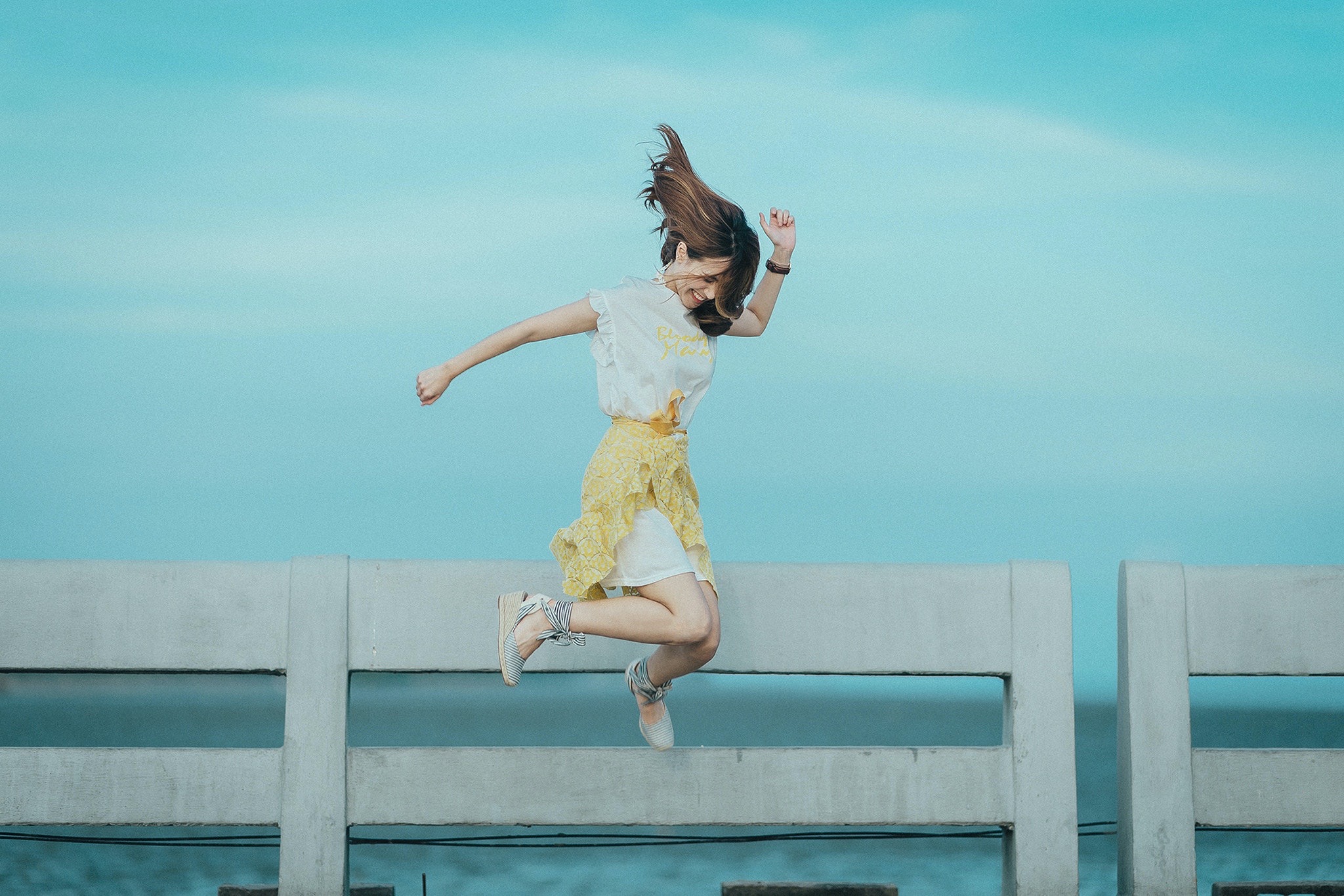 Een vrouw met een gele rok en witte blouse en lang bruin haar springt in de lucht, achter haar zie je een reling zee. Foto via pexels door: Quang Anh Ha Nguyen