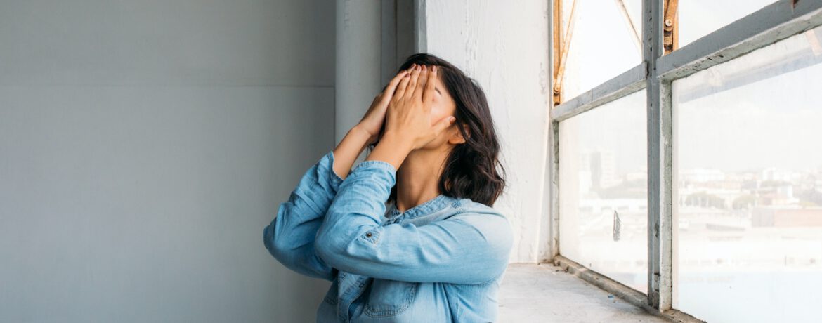 een vrouw leunt met haar rug tegen een hoge vensterbank bij een zonnig raam. Ze draagt een spijkerstof overhemd met lange mouwen en slaat beide handen voor haar gezicht, en heeft halflang sluit donker haar.