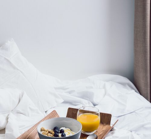 een dienblad met een kom muesli en een glas sap is geplaceerd op een beslapen maar leeg bed met wit beddengoed.