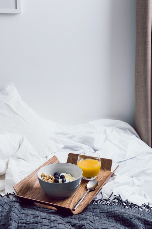 een dienblad met een kom muesli en een glas sap is geplaceerd op een beslapen maar leeg bed met wit beddengoed.